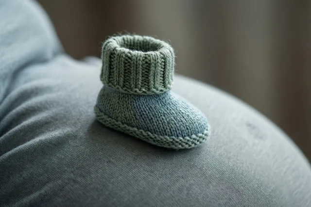 Een klein gebreid babyslofje in gedempt groen en blauw rust op een ronde zwangere buik, met zacht licht en een wazige achtergrond.