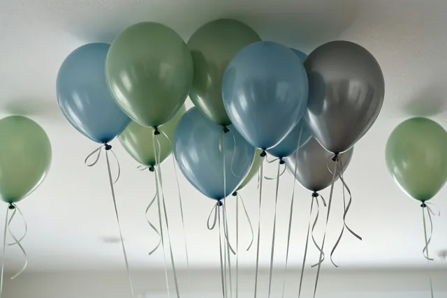 Een groep ballonnen in gedempt groen, blauw en grijs zweeft tegen een wit plafond, met lange linten naar beneden.