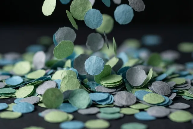 Ronde papieren confetti in groene, blauwe en grijze tinten valt tegen een donkere achtergrond.