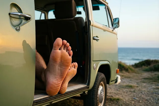 Uit de open zijdeur van een oude groene camper op een grindpad aan zee steken een grote volwassen voet en een kleinere kindervoet in goudkleurig licht.
