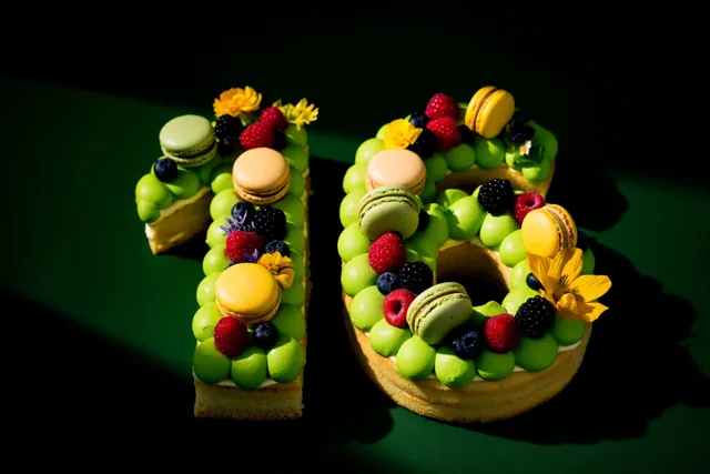 Een taart in de vorm van 16 met groen glazuur, bessen, macarons en eetbare bloemen staat tegen een donkergroen oppervlak in sterk contrasterend licht.