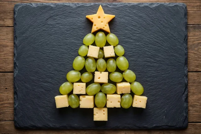 Groene druiven en gele kaasblokjes liggen in de vorm van een kerstboom met een kaasster op een donkere leistenen plank op een houten ondergrond.