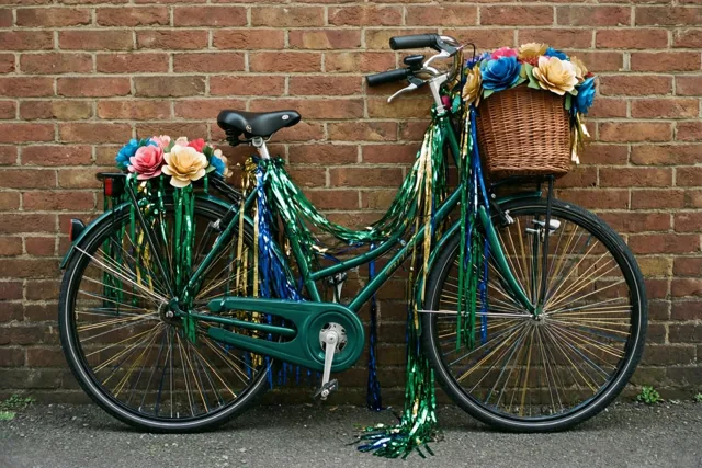 Een groene fiets met kleurrijke papieren bloemen en glanzende slingers leunt tegen een rode bakstenen muur, met groen, rood en goud als overheersende kleuren.