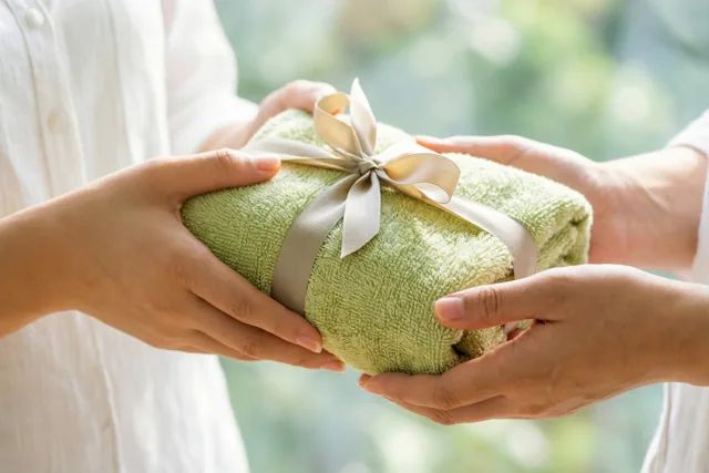Twee handen geven een netjes opgerolde groene handdoek met een beige lint door tegen een zacht onscherpe achtergrond met daglicht en groen.