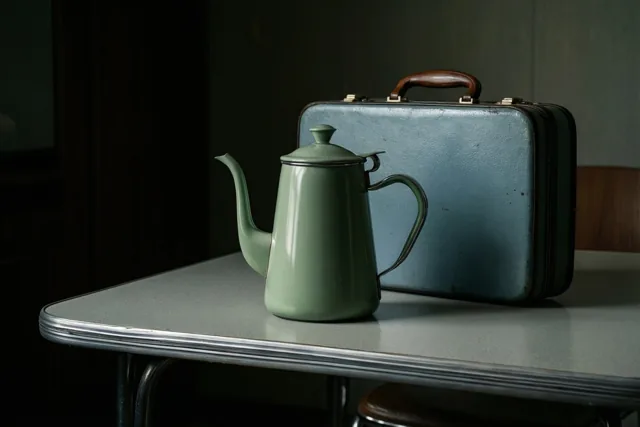 Een groene geëmailleerde koffiepot en een blauwe retroaktetas staan op een grijze formicatafel met een chromen rand in een schemerige kamer met donkere muren.