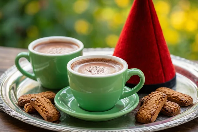 Buiten staat een zilveren dienblad met twee groene porseleinen kopjes chocolademelk en speculaasjes naast een rode mijter, tegen een wazige groene en gele achtergrond.