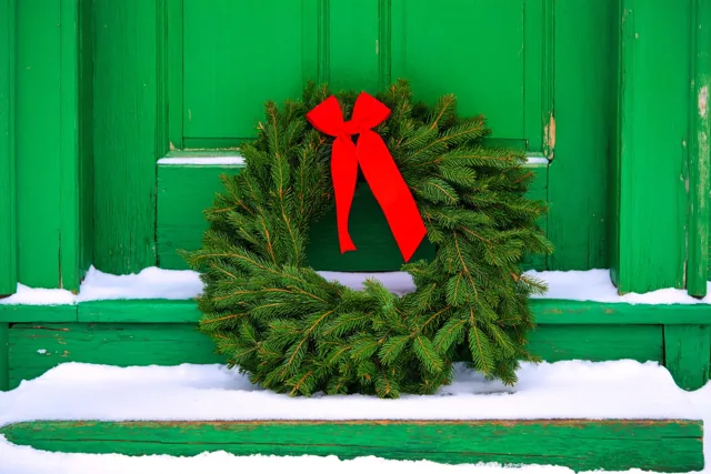 Een groene kerstkrans met een rood lint ligt op een besneeuwde trede voor een oude groene houten deur, met verse sneeuw op de treden en het deurkozijn.