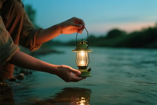 Handen houden bij schemering een brandende groene lantaarn boven een rivier, met licht dat op het water weerspiegelt in oranje en blauwe tinten.