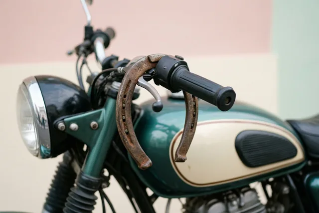 Een oude motorfiets met een groen met crèmekleurige tank heeft een roestig hoefijzer aan het stuur, tegen een zachte pastelkleurige achtergrond.