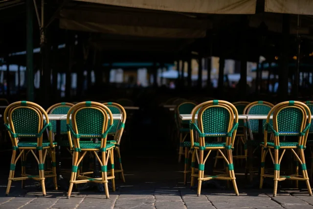 Groene stoelen en bamboestoelen staan rond witte tafels op een stenen caféterras onder een overkapping in warm zonlicht.