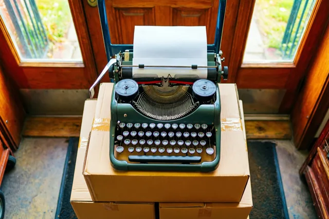 Een oude groene typemachine staat op een stapel kartonnen verhuisdozen voor een houten deur met glazen panelen, in warm daglicht.
