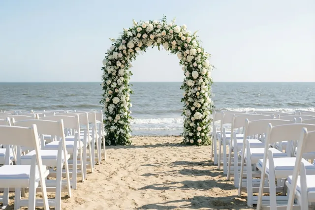 Rijen witte stoelen staan op een zandstrand tegenover een grote boog met witte en lichtroze bloemen, met de grijsblauwe zee onder een heldere hemel op de achtergrond.