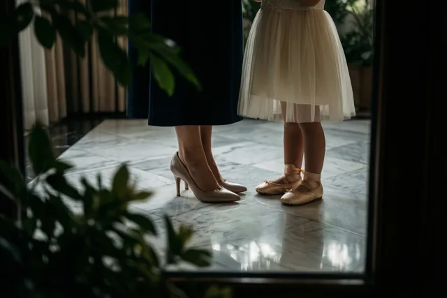 Het onderlichaam van een vrouw op elegante hakken en dat van een kind op ballerina's zijn naast elkaar te zien op een gepolijste marmeren vloer, met donker gebladerte op de voorgrond in zacht, warm licht.