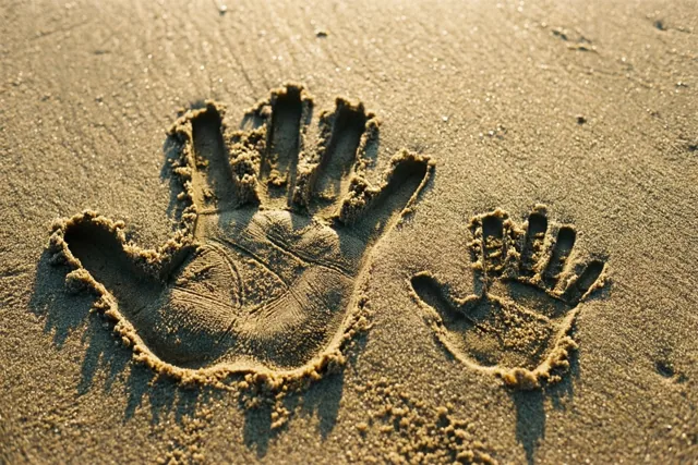Een grote en een kleine handafdruk staan naast elkaar in nat zand in warm goudkleurig licht.