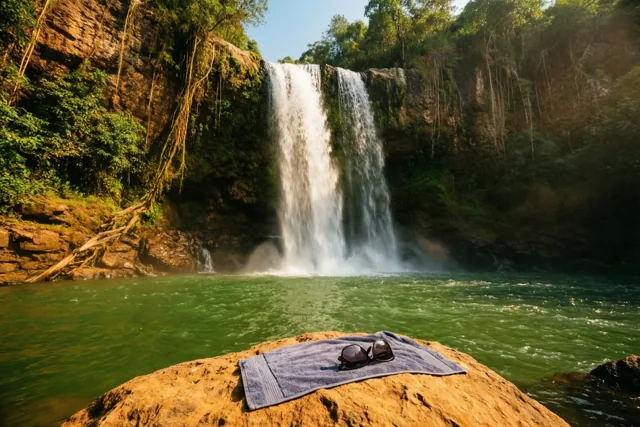 Een waterval stroomt in een groene waterpoel in een dichtbegroeide kloof, met een handdoek en zonnebril op een zonnige rots vooraan.
