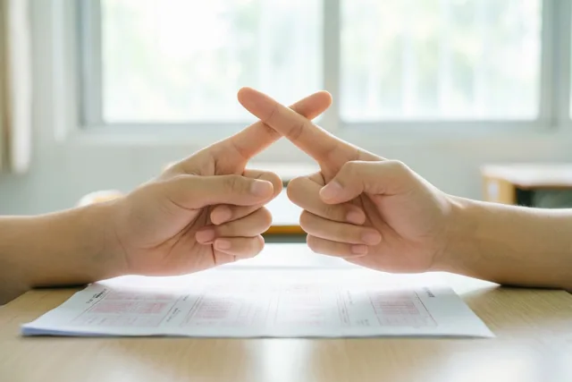 Twee handen met gekruiste vingers liggen op een houten bureau naast een omgekeerd examenblad in een helder verlichte kamer met een raam op de achtergrond.