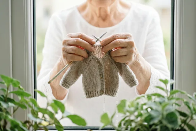 Door een raam zijn de handen van een oudere vrouw te zien die met zilverkleurige naalden een klein beige vestje breit, met groene planten op de voorgrond.