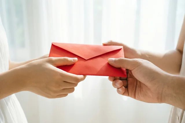 Twee paar handen geven een rode envelop door tegen een lichte, zacht verlichte achtergrond waarin rood en wit overheersen.