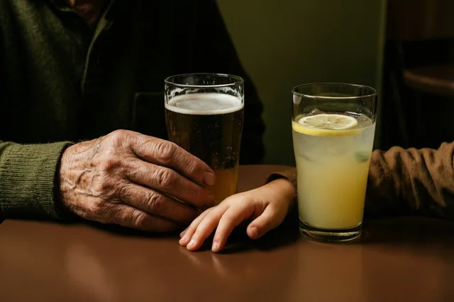 De hand van een oudere persoon en een kinderhand rusten op een bruine tafel naast een glas bier en een glas limonade in warm licht.
