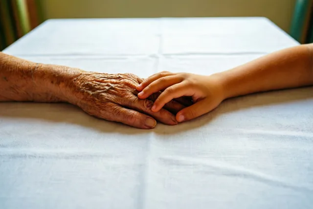 Een oudere hand en een kleine kinderhand rusten samen op een wit linnen tafelkleed in zacht licht.