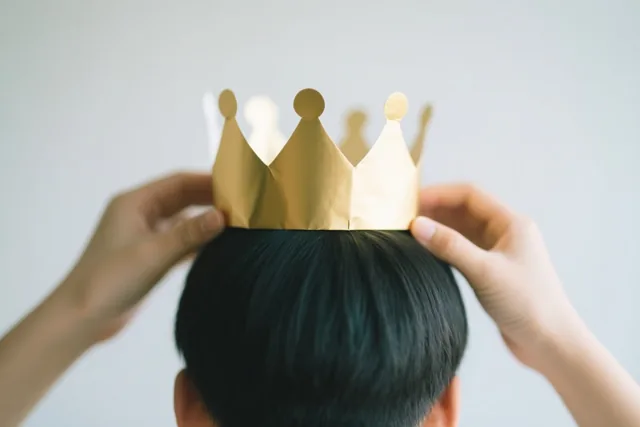 Twee handen plaatsen een goudkleurige papieren kroon op het hoofd van een persoon met donker haar, van achteren gezien tegen een lichtgrijze achtergrond.