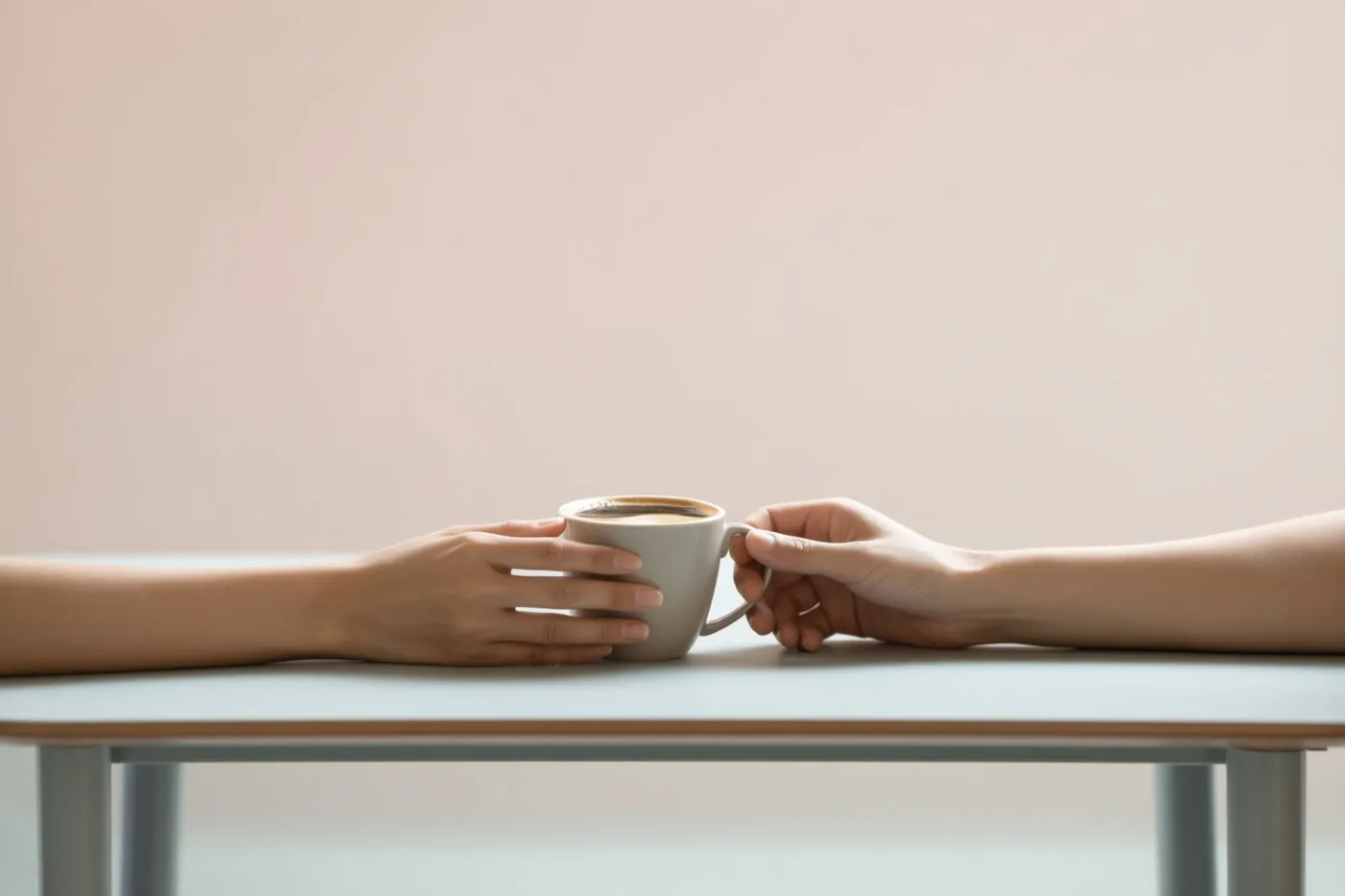 Twee handen houden samen één koffiekop vast boven een tafel tegen een effen achtergrond in zachte pasteltinten.