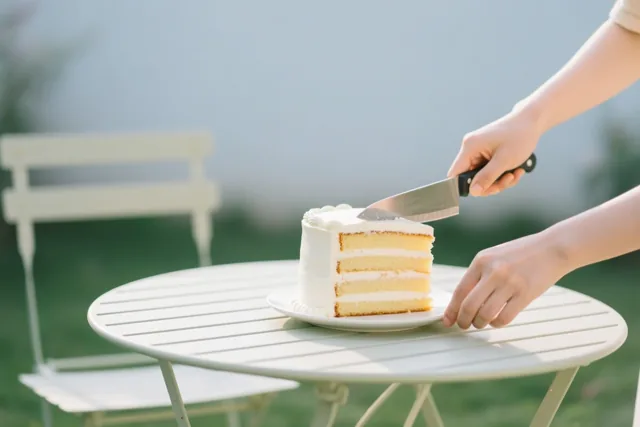 Handen snijden een stuk uit een hoge witte laagjestaart op een witte tuintafel buiten, met een lichte onscherpe achtergrond.