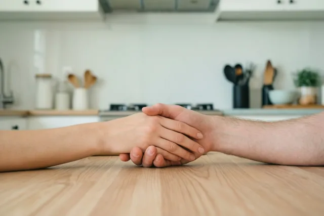 Twee handen houden elkaar vast op een houten keukentafel, voor een onscherpe moderne keuken met witte kasten en houten keukengerei.