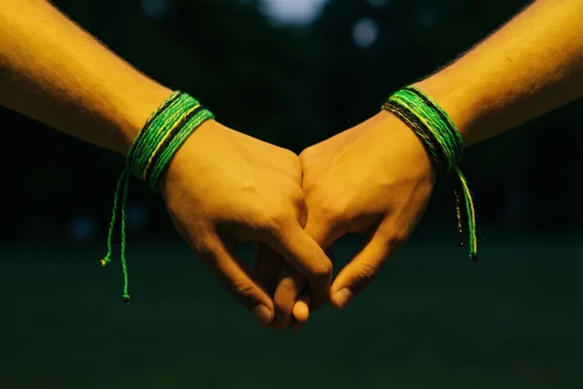 Twee handen met dezelfde groene en gele vriendschapsarmbandjes vormen samen een hart in warm avondlicht tegen een donkere, onscherpe achtergrond.