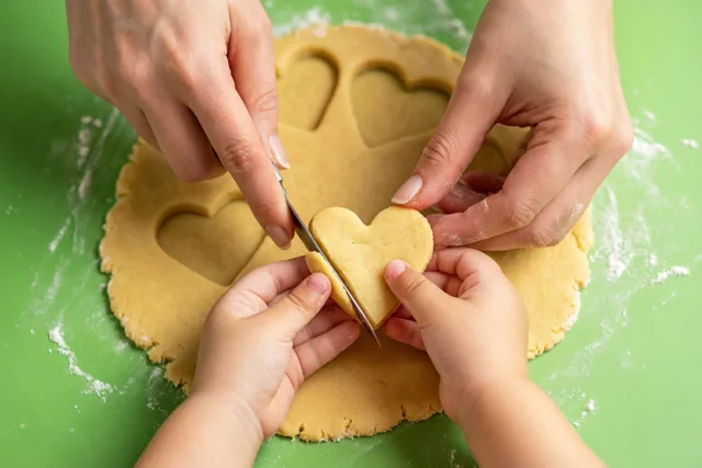 Een volwassene en een kind steken met hun handen hartvormige koekjes uit uitgerold deeg op een groene ondergrond.