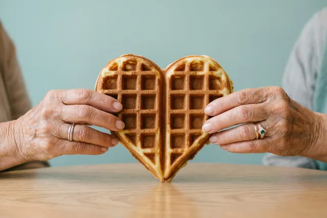 Twee oudere handen met trouwringen houden een hartvormige wafel vast bij een houten tafel tegen een zachtblauwe achtergrond.