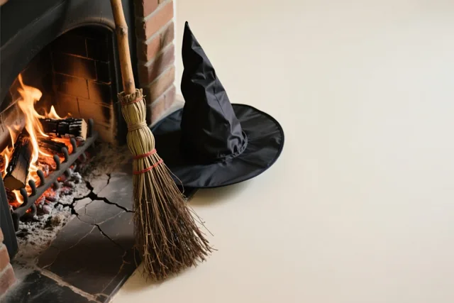 Een zwarte puntige heksenhoed en een rustieke bezem staan naast een open haard met brandende vlammen in een warm verlichte kamer.