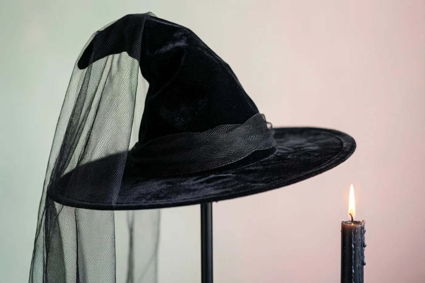 Een zwarte fluwelen heksenhoed met een zwarte sluier staat op een standaard naast een brandende zwarte kaars tegen een achtergrond met een zacht kleurverloop in gedempte tinten.
