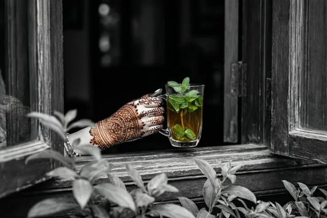 Een hand met fijne hennapatronen houdt een glazen mok muntthee op een houten vensterbank vast, met alleen de groene munt en amberkleurige thee in kleur.