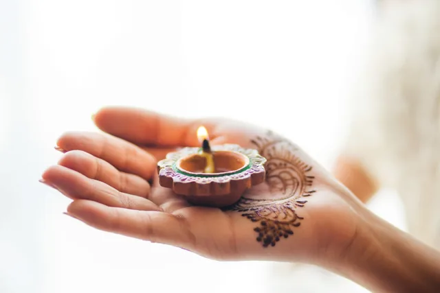 Een hand met fijne hennapatronen houdt een brandend lampje van klei vast tegen een lichte, onscherpe achtergrond in gouden en aardse tinten.