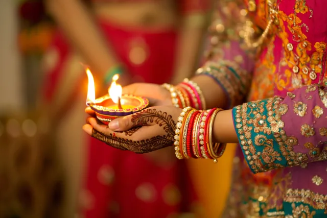 Van dichtbij zijn handen met fijne hennapatronen en een brandend olielampje te zien bij traditionele kleding in roze, paars en blauwgroen.