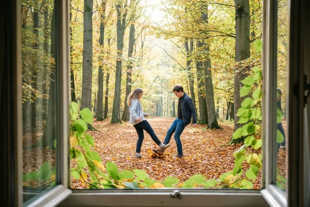 Door een open raam zie je twee vrienden tegen herfstbladeren schoppen in een beukenbos, met groene bladeren op de voorgrond.