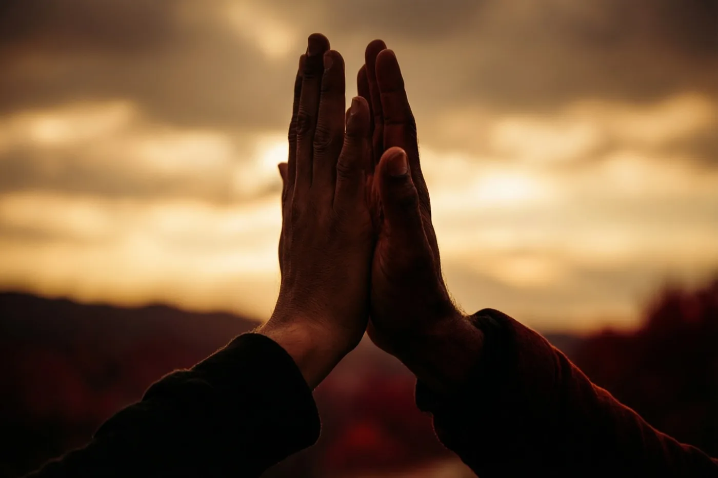 Twee handen geven elkaar buiten een high five tegen een goudkleurige hemel bij zonsondergang, met oranje, gele en donkerbruine tinten.