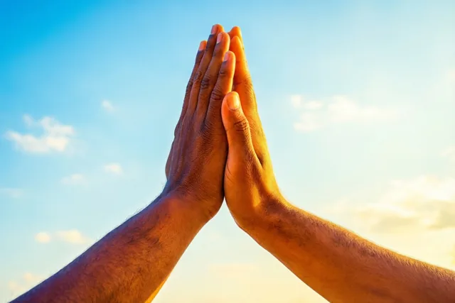 Twee handen raken elkaar in een high five tegen een helderblauwe hemel met verspreide wolken, in warm zonlicht.