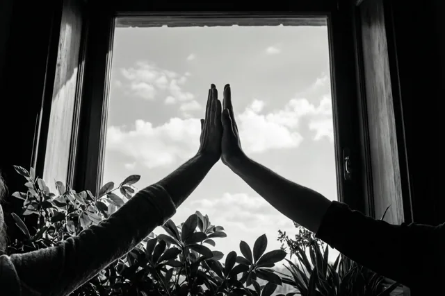 Twee handen geven elkaar een high five voor een raam met een heldere hemel, in zwart-wit met donkere silhouetten tegen het licht.