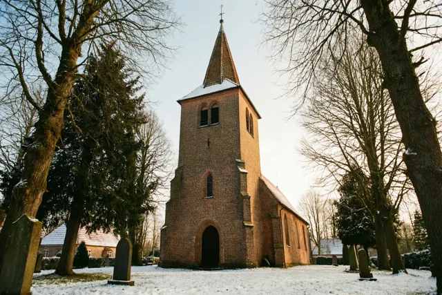 Een kleine bakstenen kerk met een hoge toren en een spits dak staat op een besneeuwd kerkhof tussen kale bomen in zacht winterlicht.