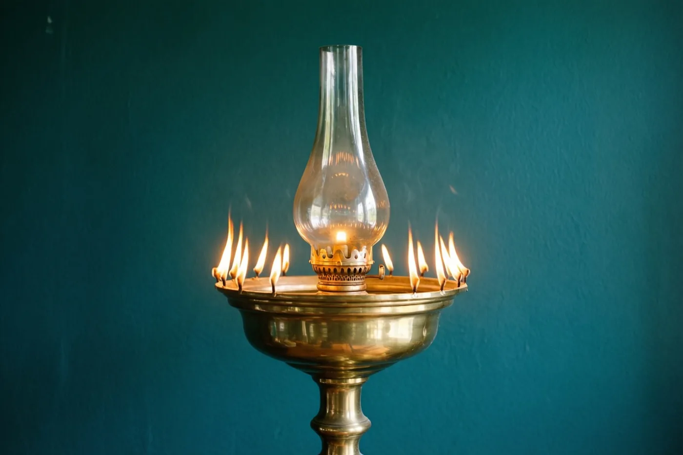 Een hoge messing olielamp met meerdere brandende lonten staat voor een donkere blauwgroene muur.