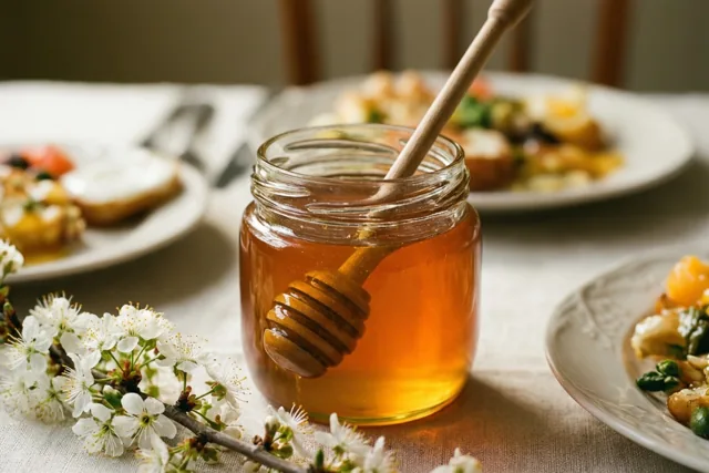 Een glazen pot goudkleurige honing met een houten honinglepel staat tussen witte bloesemtakken op een zacht verlichte ontbijttafel, met borden eten op de achtergrond.