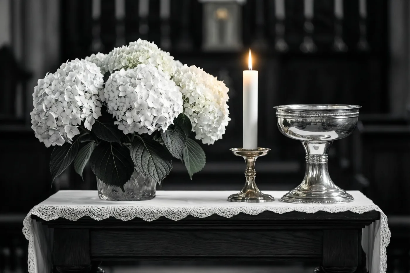 Op een klein altaar staan witte hortensia's in een glazen vaas, een brandende doopkaars in een messing houder en een zilveren doopvont tegen een donkere achtergrond.