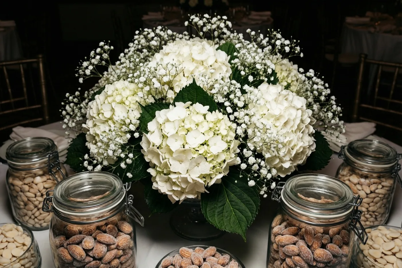 Een tafelstuk met grote witte hortensia's en gipskruid staat tussen glazen potjes suikerbonen op een wit tafelkleed, met donkere stoelen op de achtergrond.
