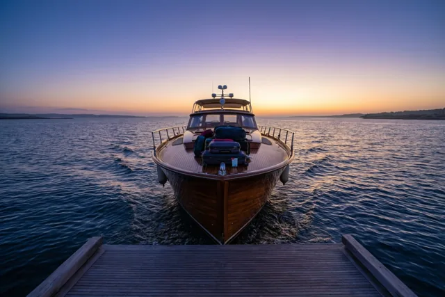Een slanke houten boot met een ingepakte koffer op het dek vaart bij zonsopgang weg van een houten pier op een kalme zee onder een oranje-paarse hemel.