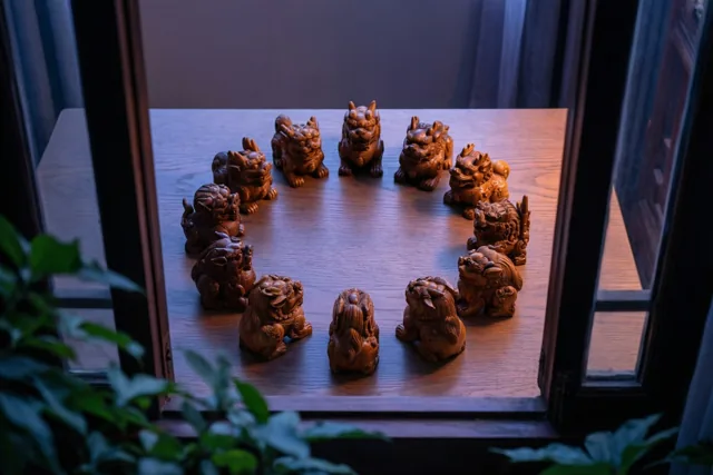 Door een raam zijn twaalf houten dierenbeeldjes van de Chinese dierenriem in een kring op een houten tafel te zien, met gedempt warm licht en groen op de voorgrond.