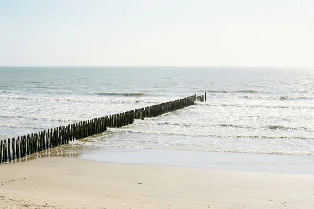 Een lange houten golfbreker loopt van een zandstrand de zee in, met zachte golven onder een bleke hemel in beige, grijze en blauwe tinten.