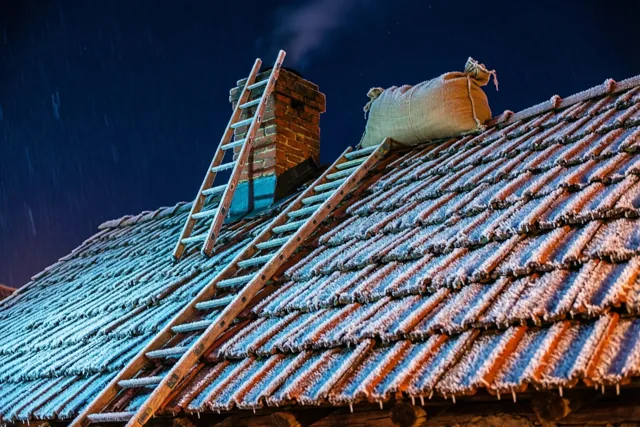 Een houten ladder leunt tegen een bakstenen schoorsteen op een berijpt pannendak, met een jutezak op de nok in blauw en oranje kunstlicht.