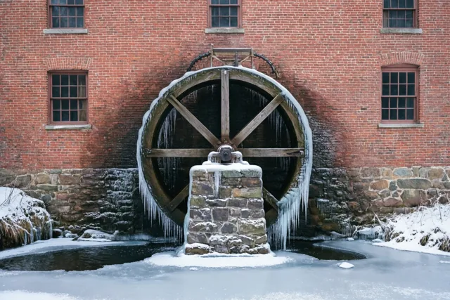 Een groot houten waterrad met ijspegels staat bij een bevroren beek voor een oude rode bakstenen molen, met sneeuw op de grond.